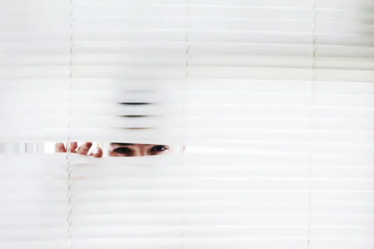 Close-up of a person's eyes peeking through parted white venetian blinds representing the hidden fees in loans that aren't always easy to spot.