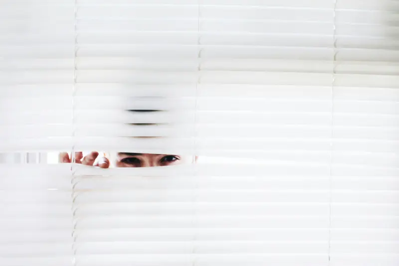 Close-up of a person's eyes peeking through parted white venetian blinds representing the hidden fees in loans that aren't always easy to spot.
