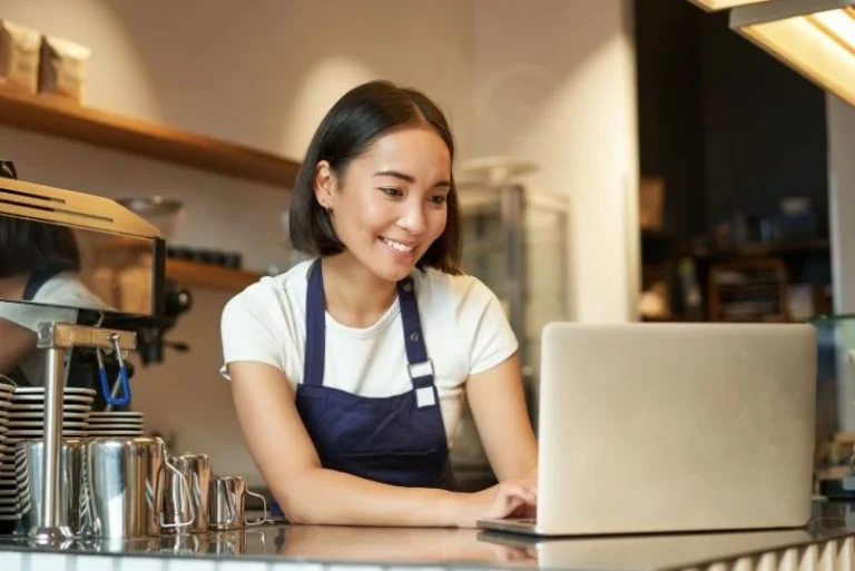 Smiling female barista in a navy apron working on a laptop behind a cafe counter, a small business owner choosing between low doc vs standard loans.