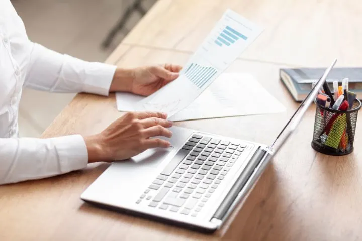 Woman in white shirt holding a printed bar chart report while using a laptop on a wooden desk preparing financial statements for review.