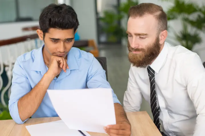 Man in blue shirt listening intently while a suited advisor in a tie reviews a document at a desk knowing what to ask before you sign matters.