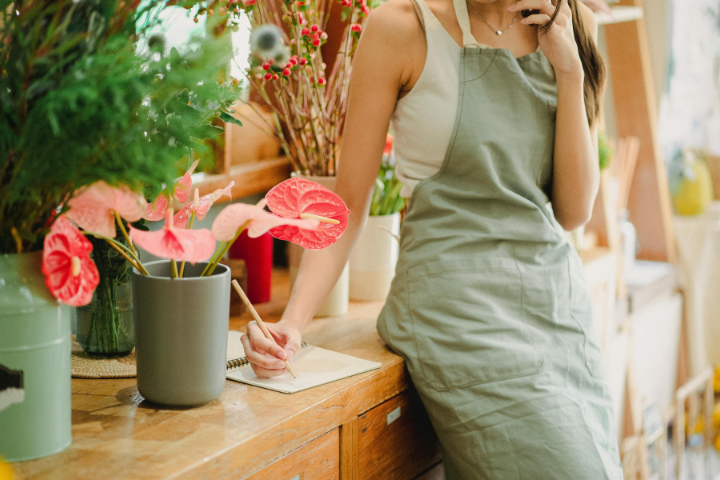 Woman in a green apron writing in a notebook while on a phone call in a flower shop running a self-employed business.