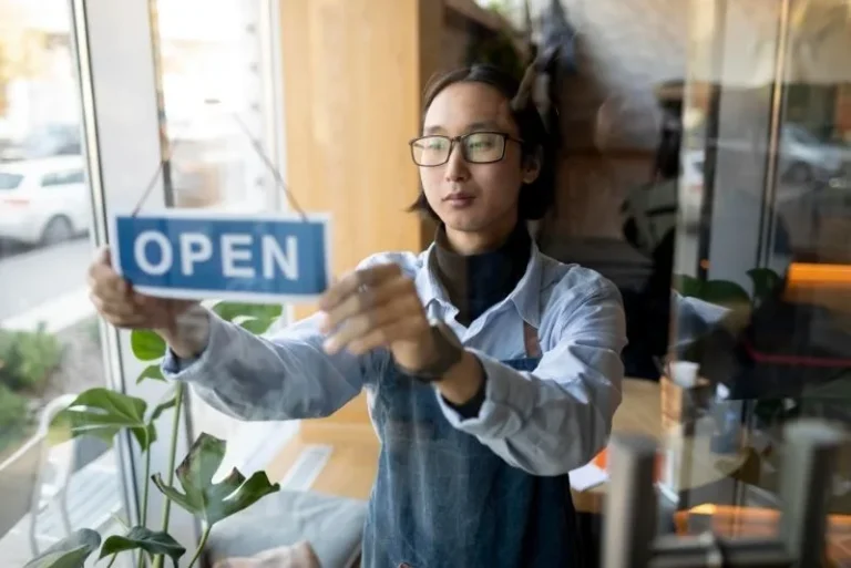 Young man in glasses and denim apron hanging an Open sign on a glass shop door, a newly established business opening for the first time.