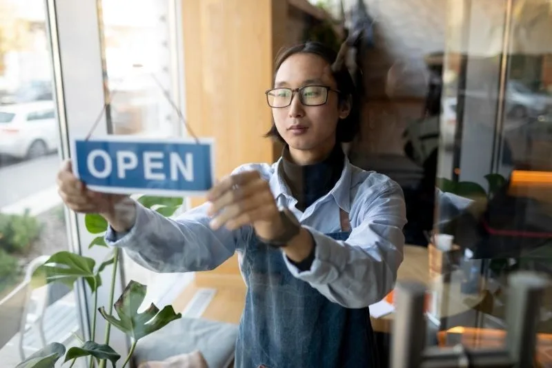 Young man in glasses and denim apron hanging an Open sign on a glass shop door, a newly established business opening for the first time.