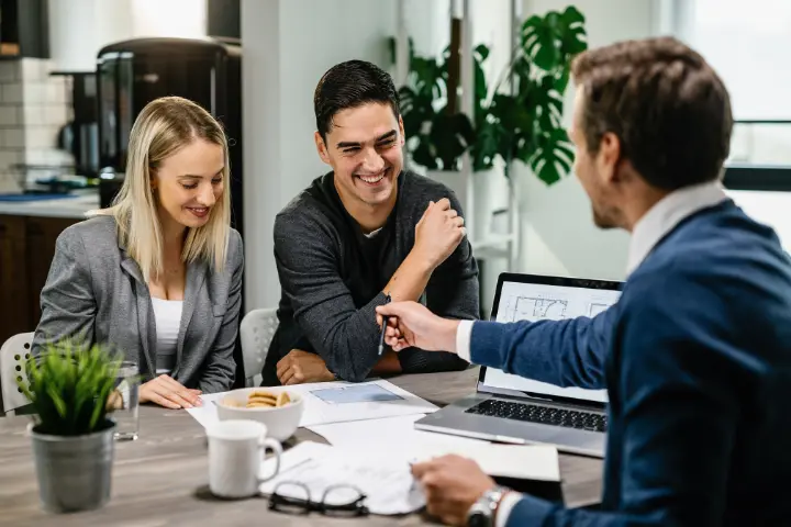 Couple smiling across a table from a suited advisor with documents, a laptop, and coffee mugs during a finance consultation.